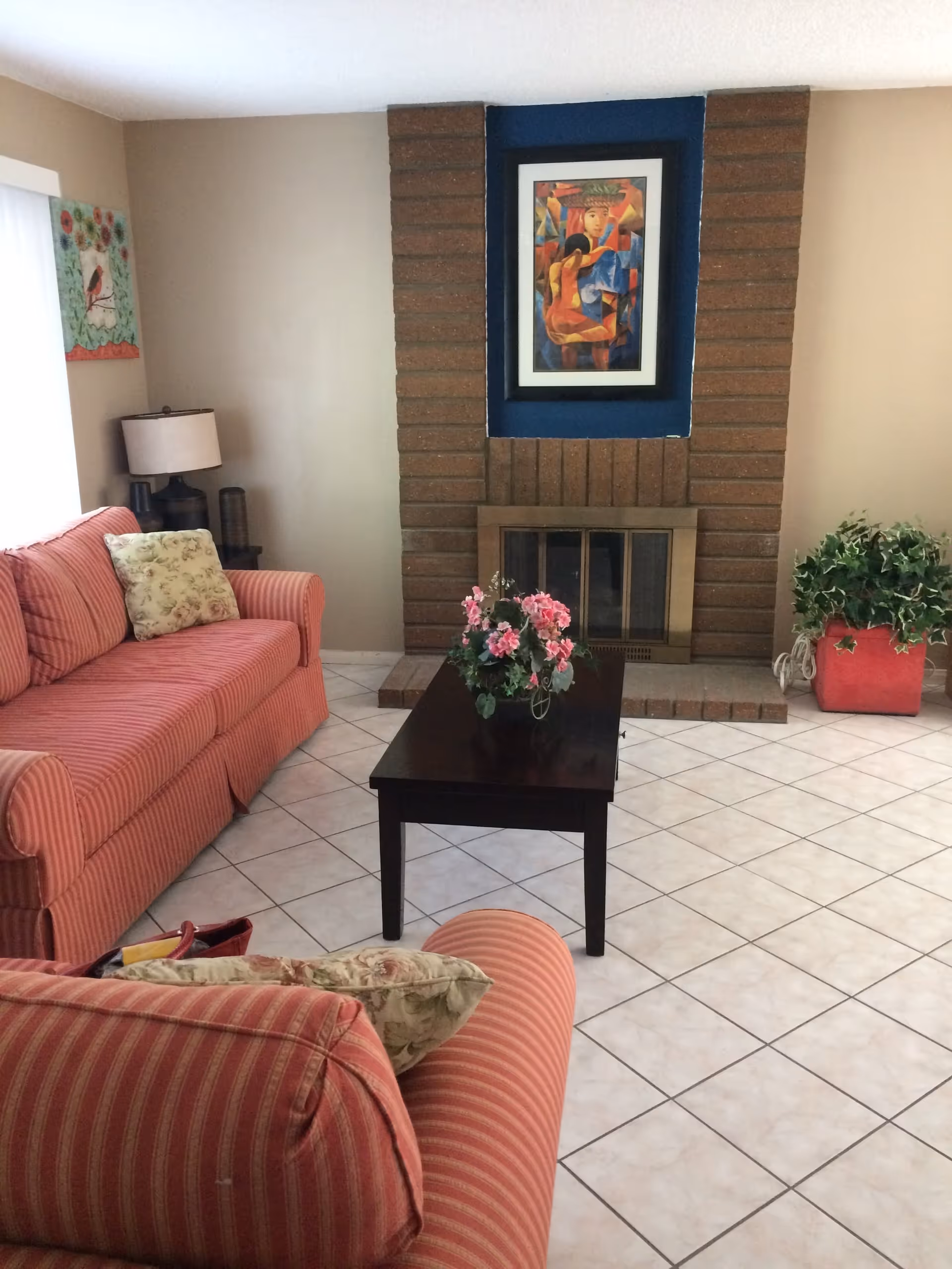 Cozy living room with striped pink sofas, a dark wood coffee table topped with a floral arrangement, and a brick fireplace with framed artwork above it.