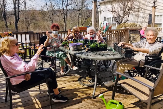 Five elderly women sitting around a metal outdoor table on a wooden deck, engaging in gardening activities with potted plants and soil trays. Some women are in wheelchairs, and one is wearing a face mask. The background shows leafless trees and part of a building under a clear sky.