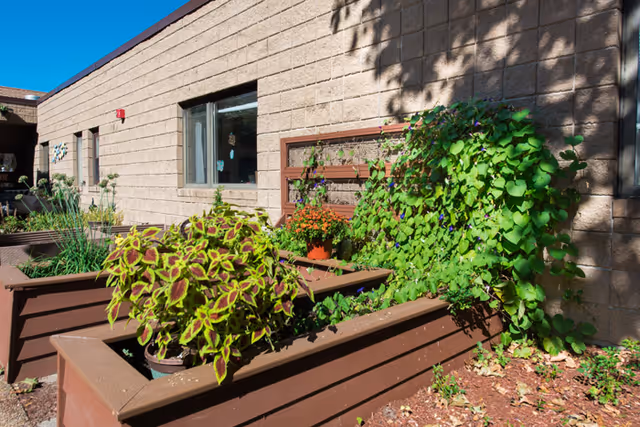 Outdoor garden area at St. Teresa Rehabilitation and Nursing Center featuring raised brown wooden planters filled with various green plants and flowers, set against a beige brick building under a clear blue sky.