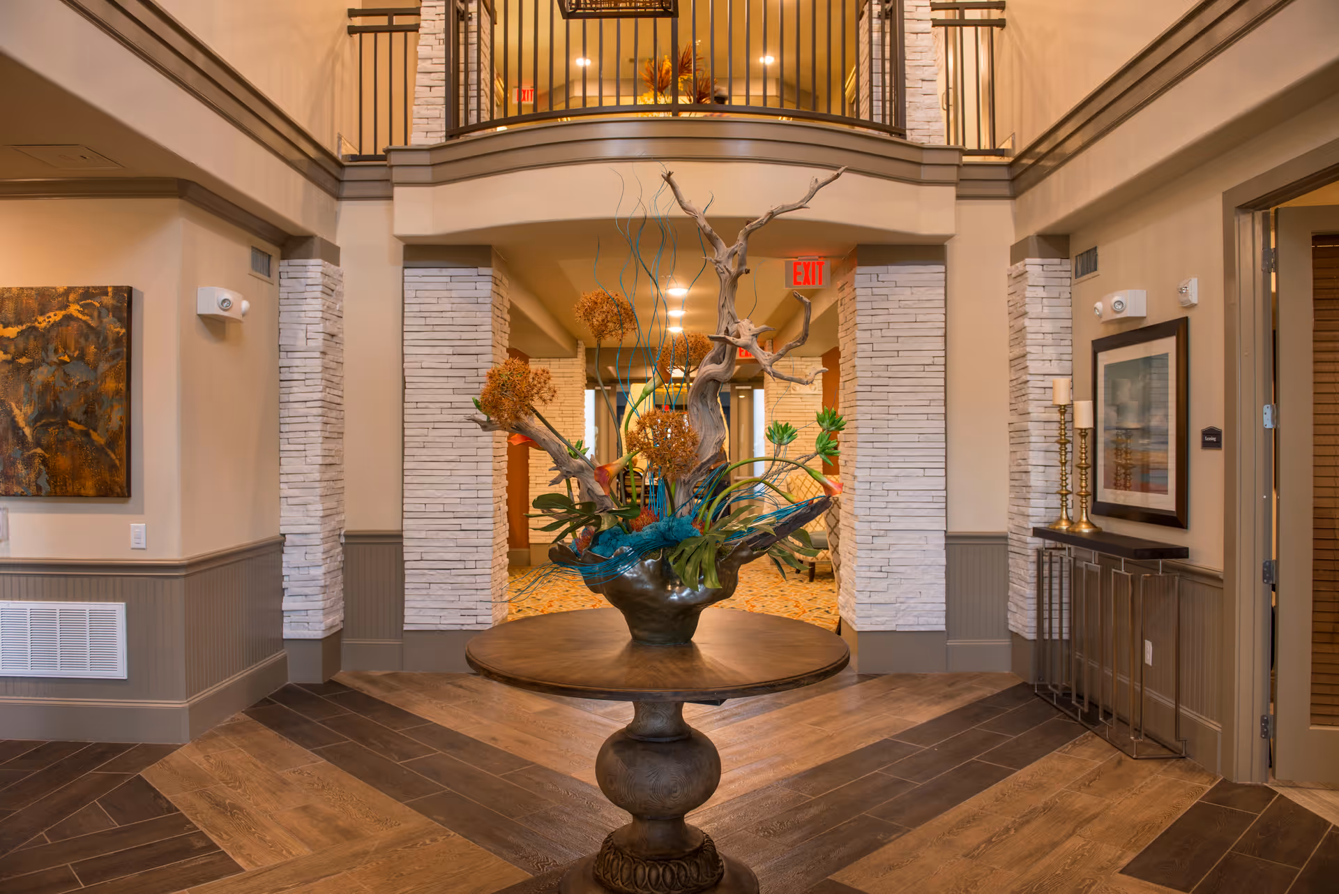 Interior view of a senior living facility lobby with a round wooden table in the center holding a decorative floral arrangement. The space features beige walls with white brick accents, a two-story ceiling with a balcony railing above, and warm lighting. There are framed artworks and candle holders on the walls, and an exit sign is visible in the background.