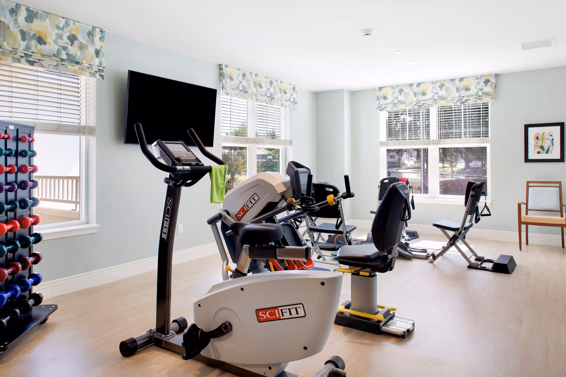 A bright exercise room with multiple pieces of fitness equipment including stationary bikes and weight machines. There is a rack of colorful dumbbells on the left side, a large flat-screen TV mounted on the wall, and windows with patterned valances letting in natural light. A wooden chair with a cushion is placed near the window on the right.