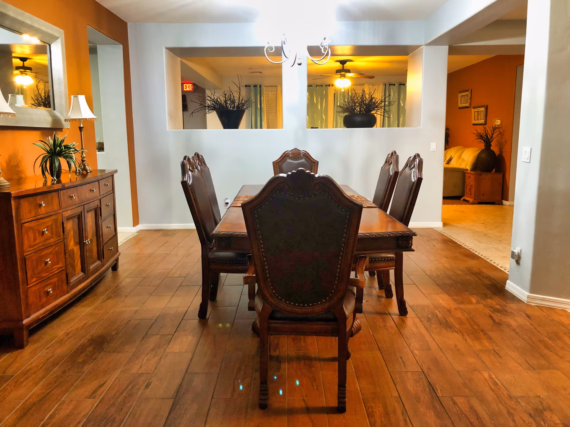A dining room with a wooden dining table and six ornate chairs. The room has wooden flooring and a sideboard with plants and lamps on it. There are two large rectangular openings in the wall, showing a glimpse of another room with ceiling fans and plants. The walls are painted in warm tones with some decorative elements.