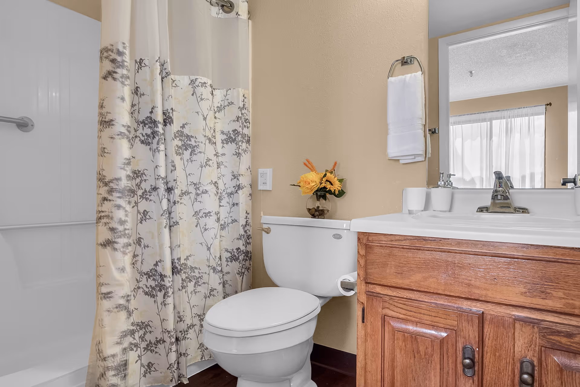 A bathroom with a white toilet, a wooden vanity with a white sink, a mirror above the sink, a towel hanging on a ring, and a shower with a floral patterned curtain. A small vase with yellow flowers is placed on the toilet tank.