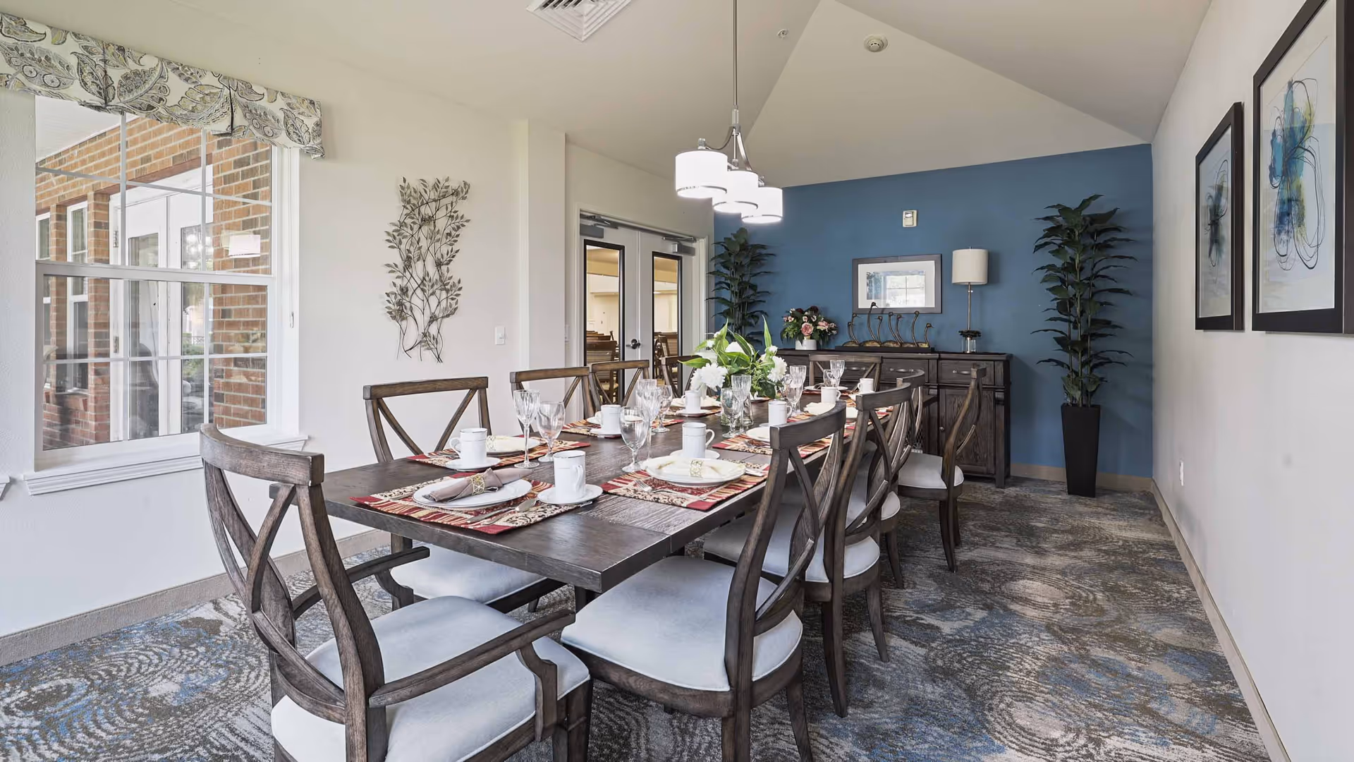 A well-lit dining room with a long wooden table set for eight people with plates, glasses, and napkins. The room features a blue accent wall with a sideboard, decorative plants, and framed artwork. A large window on the left side lets in natural light, and a modern chandelier hangs above the table.