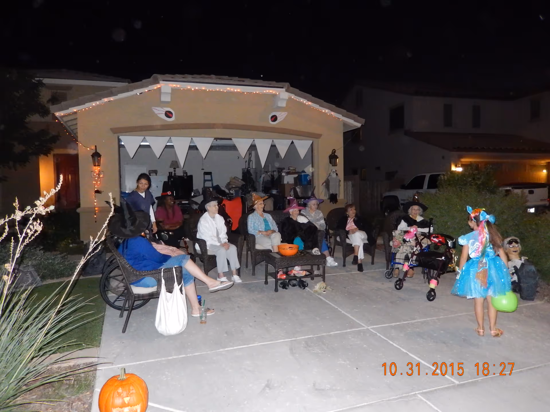 A group of elderly people sitting outside a house decorated with Halloween decorations, including a pumpkin and a banner resembling teeth. Some are wearing witch hats and costumes. A young child in a blue costume is standing nearby holding a green bucket, likely for trick-or-treating. It is nighttime.