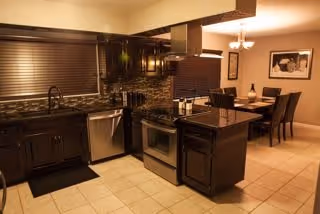 Modern kitchen with dark wood cabinets, a central island with a black countertop, stainless steel appliances including a dishwasher and stove, and a dining area with a table and chairs in the background.
