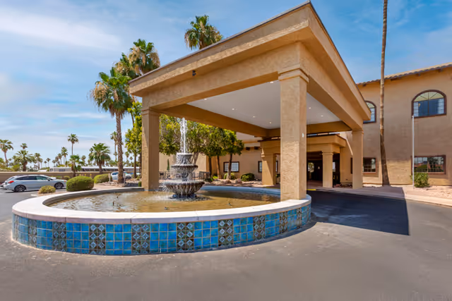 Entrance of Brookdale Springs Mesa facility featuring a large covered driveway with a tiered water fountain in the center, surrounded by palm trees and parked cars under a clear blue sky.