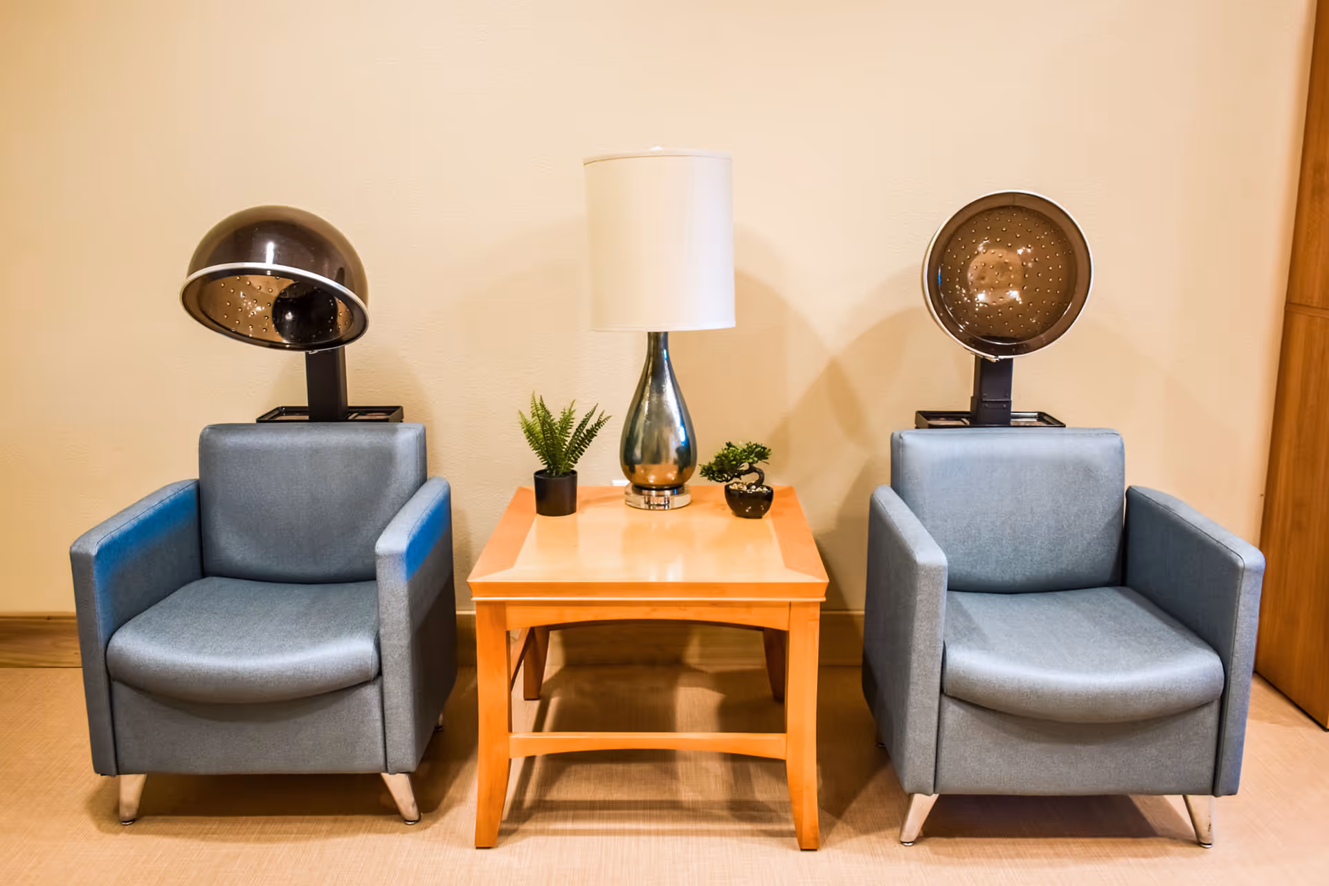 Two blue salon chairs with hair drying hoods attached, positioned on either side of a wooden side table with a lamp and two small potted plants, set against a beige wall.