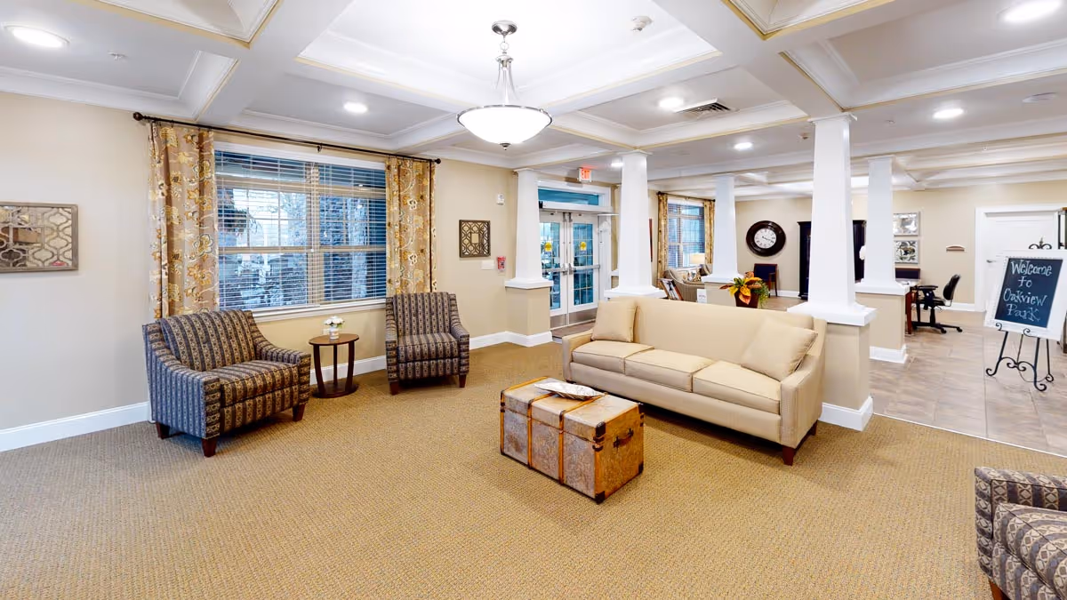 A bright and spacious living room area in Oakview Park facility featuring a beige sofa, two patterned armchairs, a vintage trunk used as a coffee table, large windows with floral curtains, and a welcoming sign near the entrance.