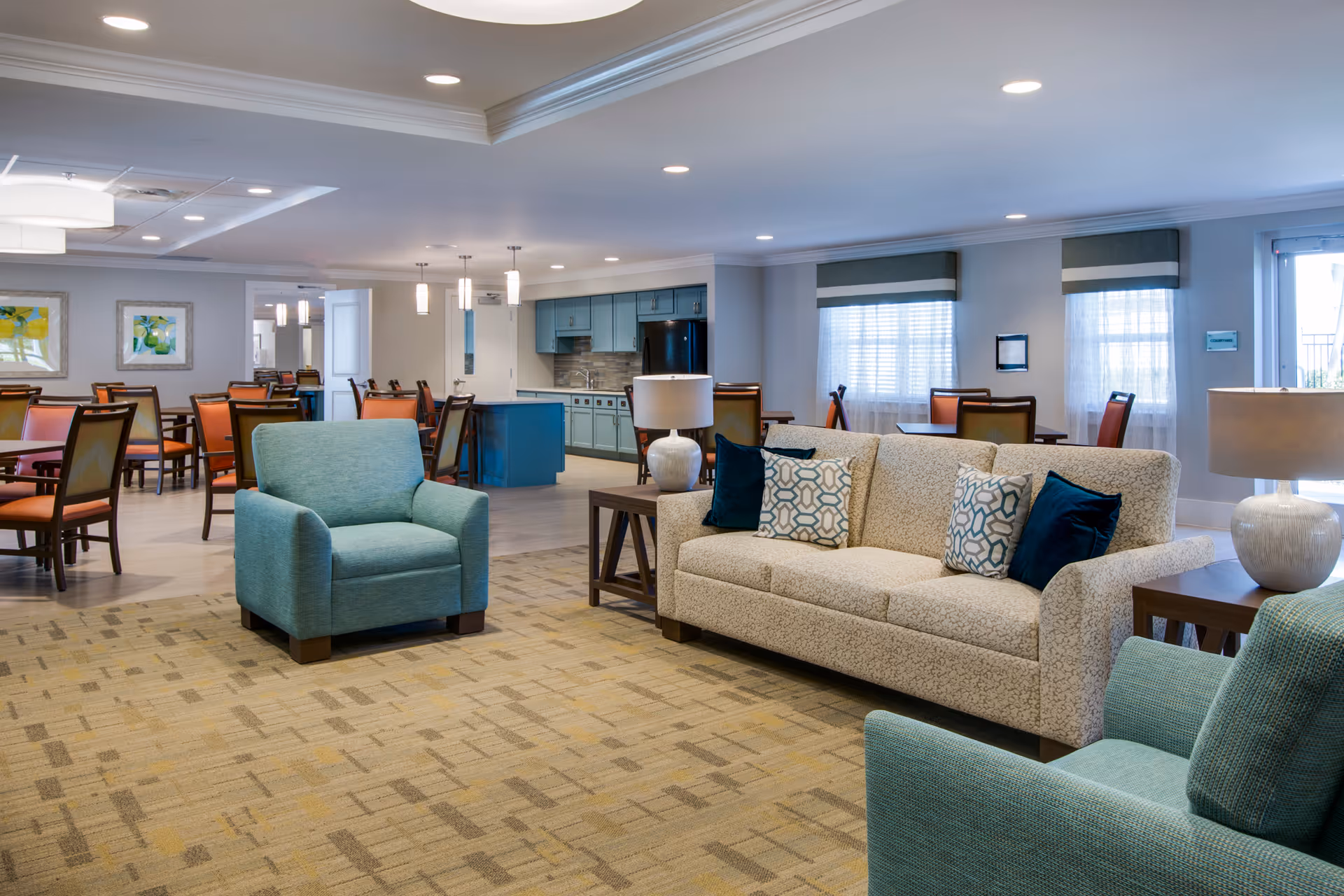 A spacious and well-lit common area in a senior living facility featuring a beige sofa with patterned and solid blue cushions, two teal armchairs, wooden side tables with lamps, and a dining area with multiple tables and chairs in the background. The room has large windows with sheer curtains and a kitchen area with blue cabinetry and modern lighting fixtures.
