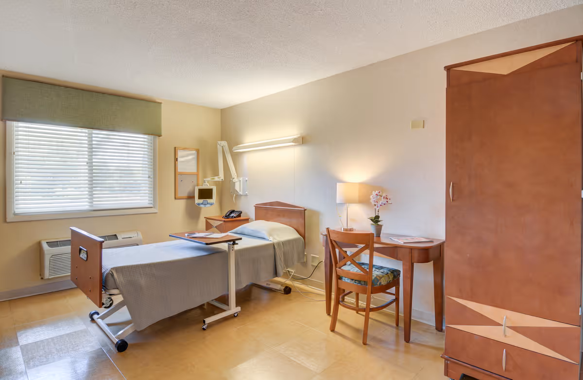 A clean and well-lit patient room in a senior living facility featuring a single hospital bed with gray bedding, an adjustable overbed table, a wooden desk with a chair, a lamp, and a potted orchid. The room has a large window with blinds and a green valance, a wall-mounted medical monitor, and a wooden wardrobe.