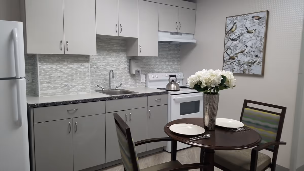 A small kitchen area with light gray cabinets, a white refrigerator, a white stove with a kettle on it, and a sink with a modern faucet. There is a round wooden table set for two with white plates, silverware, and a vase of white flowers. A framed artwork featuring birds hangs on the wall behind the table.