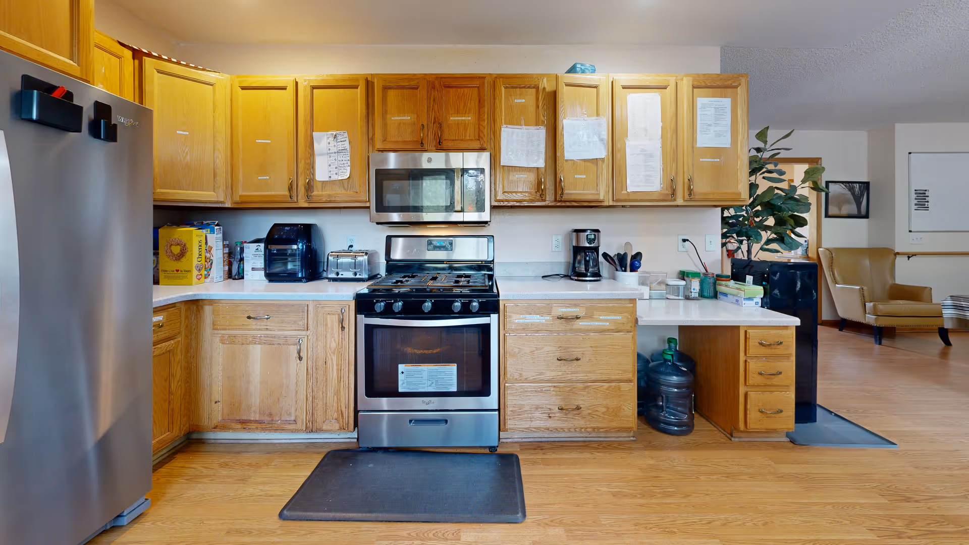 A kitchen area with wooden cabinets and drawers, a stainless steel refrigerator on the left, a stainless steel stove with an oven and microwave above it in the center, and a coffee maker on the right countertop. There are various kitchen appliances and items on the countertops, including a toaster, an air fryer, and a water jug under the counter. The floor is wooden, and part of a living area with chairs and a plant is visible in the background.