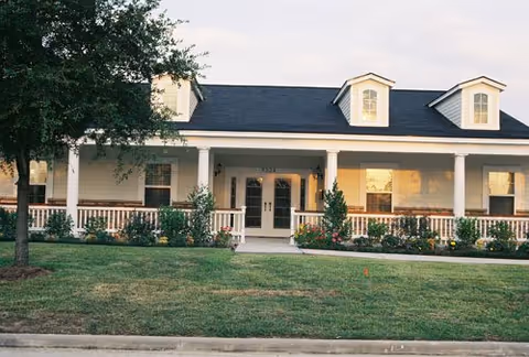 Front exterior view of a single-story building with a covered porch, white columns, and dormer windows on the roof. The building is surrounded by a well-maintained lawn, flower beds, and a tree on the left side.