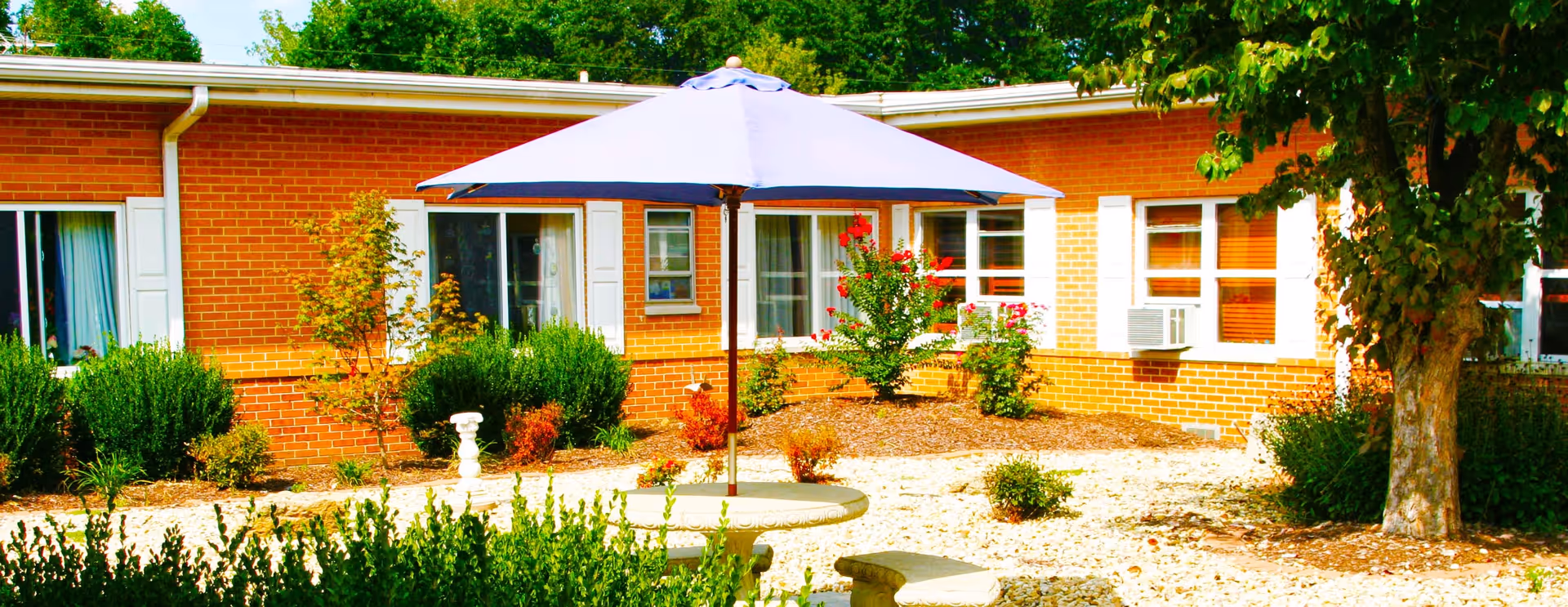 Outdoor courtyard area of a brick building with several windows, surrounded by green bushes, small trees, and plants. In the center, there is a round concrete table with a large blue umbrella providing shade, and curved concrete benches around the table. The ground is covered with light-colored gravel and mulch.