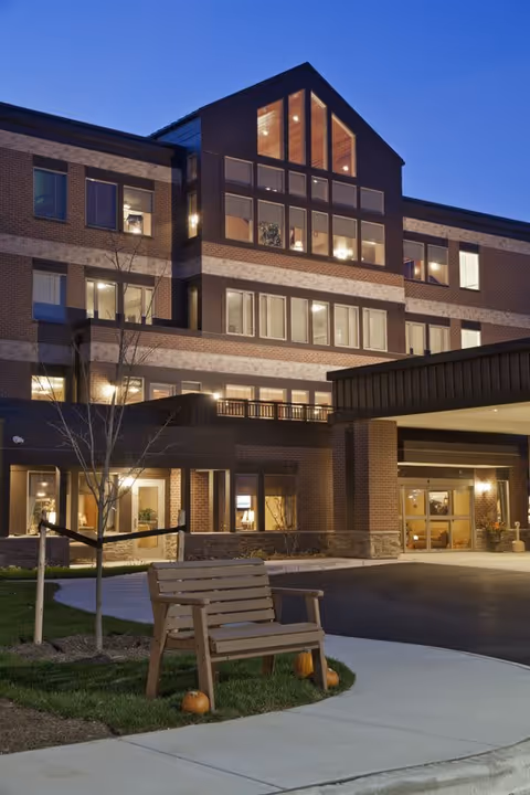 Exterior view of a multi-story senior living facility building at dusk with lights on inside. The building features large windows and a covered entrance. In the foreground, there is a wooden bench on a small grassy area with three small pumpkins placed around it, and a young tree supported by stakes.