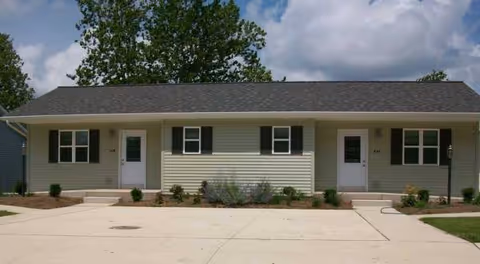 Single-story building front with two doors, light siding, and shrubs along the walkway.