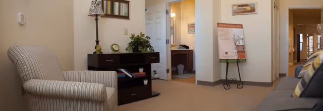 Interior seating area with an upholstered armchair and sofa, a console table with lamp and plants, and open doorways to adjacent rooms.