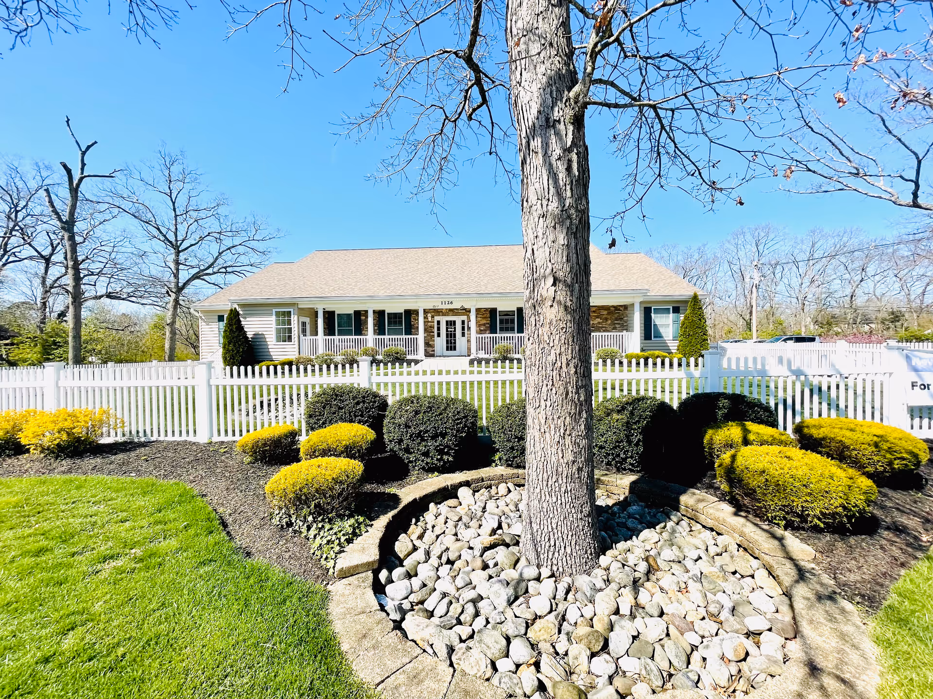 A single-story building with a beige roof and white siding, surrounded by a white picket fence. In front of the fence is a landscaped garden with trimmed bushes, yellow shrubs, and a tree planted in a circular stone-bordered bed filled with rocks. The sky is clear and blue.