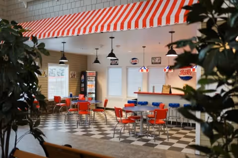 Interior view of a retro-style dining area with red and white striped awning, red chairs, blue bar stools, checkered black and white floor, pendant lights, and vintage soda signs on the walls.