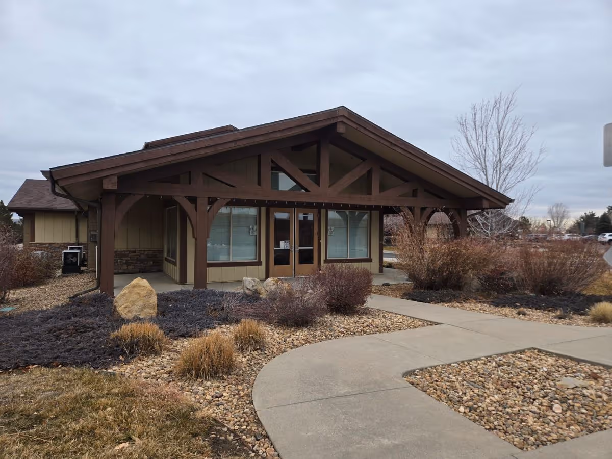 Single-story senior living facility entrance with a gabled wooden porch, landscaped rock beds, and a curved concrete walkway.