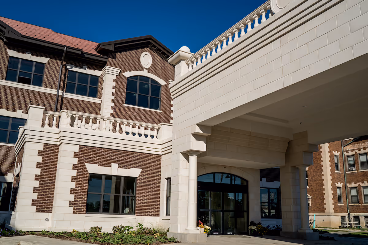 Exterior view of Meadowbrook Manor - Lagrange, showing a brick building with white stone accents, large windows, and a covered entrance supported by columns under a clear blue sky.