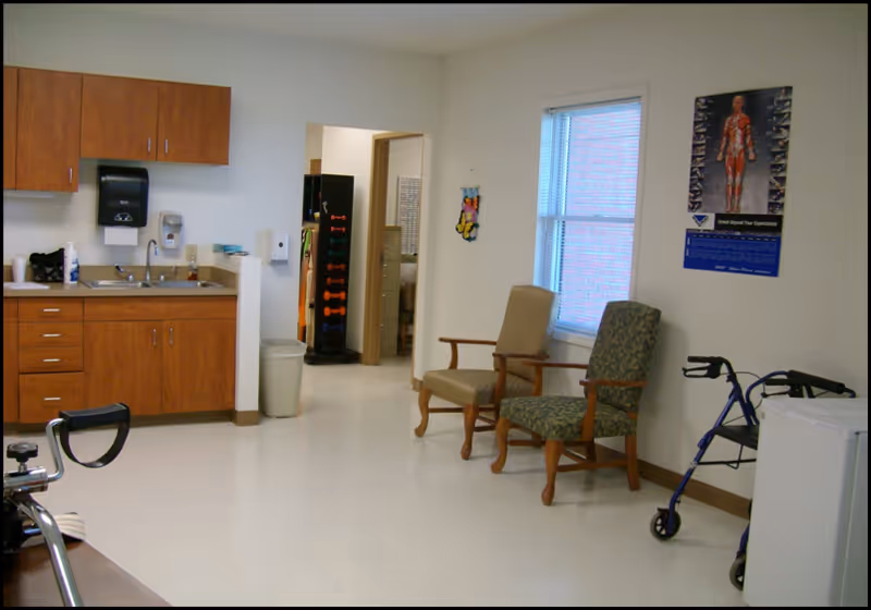A room with a small kitchenette area featuring wooden cabinets, a sink, and a paper towel dispenser. Two upholstered chairs are placed near a window with blinds. A walker is positioned next to a small white refrigerator. A poster showing a human muscular system is on the wall. The floor is light-colored and the walls are white.