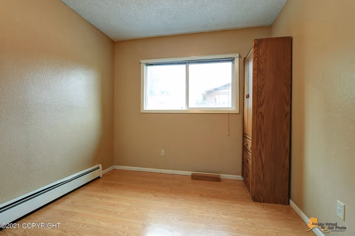 Small empty bedroom with wood floors, a window, baseboard heater and a tall wooden cabinet.