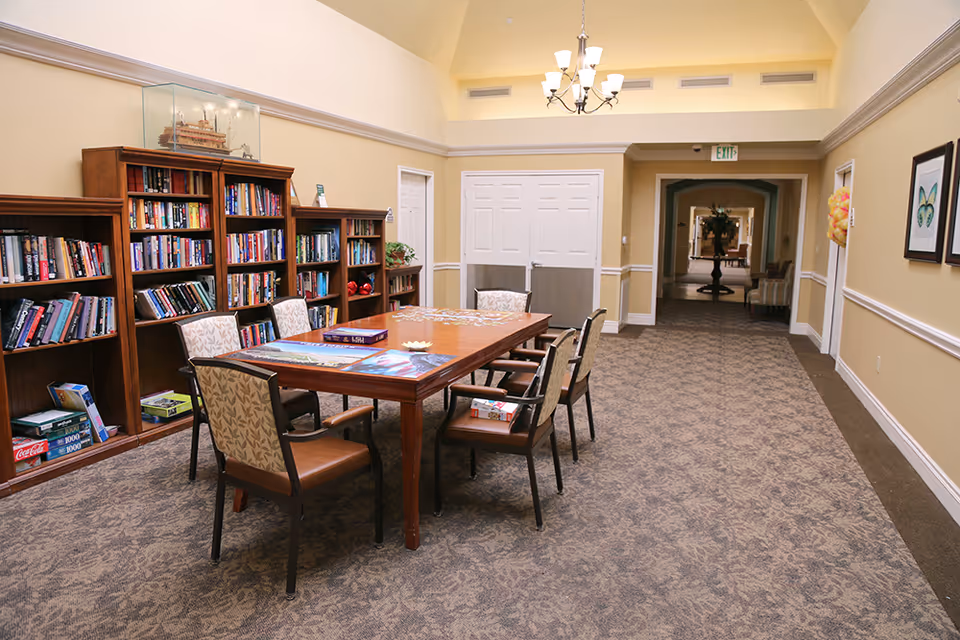 A bright interior common room with a large wooden table and chairs, bookshelves along the left wall, and a hallway leading deeper into the facility.