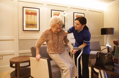 A caregiver in a blue uniform assists an elderly woman with a walker as she stands up from a chair in a well-lit room with framed artwork on the walls and comfortable seating.
