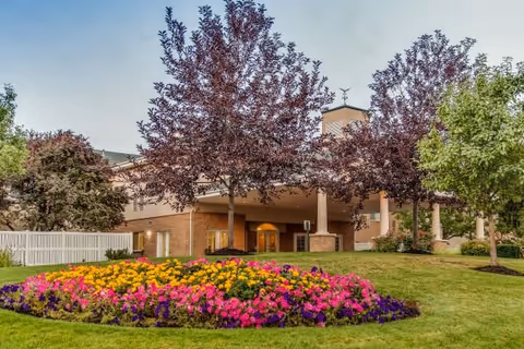 A landscaped garden area in front of a building with a flower bed featuring vibrant pink, yellow, and purple flowers. Several trees with dark red and green foliage are planted around the garden, and the building has a covered entrance with columns and a cupola on the roof.