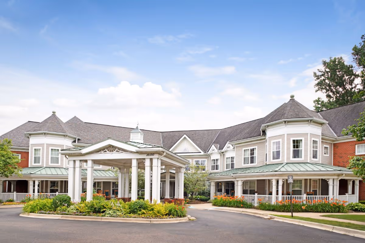 Two-story senior living building front with a covered porte-cochere, wraparound porch, and landscaped circular driveway under a blue sky.