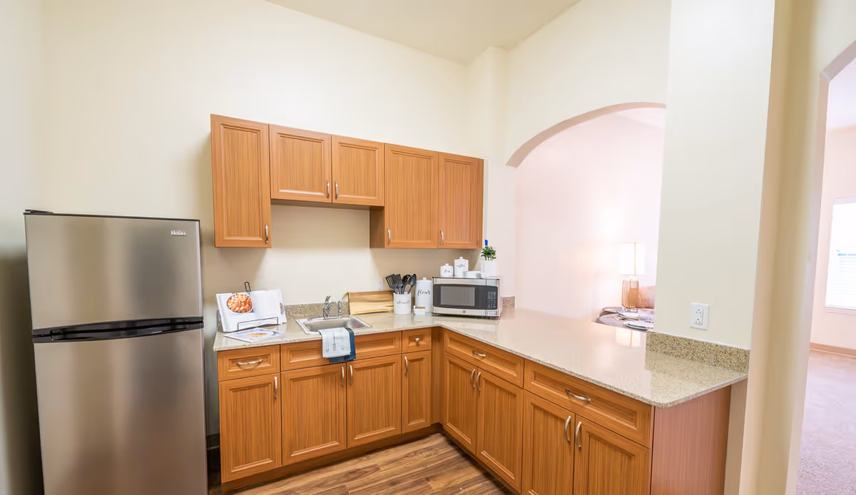 A small kitchen area with wooden cabinets, a stainless steel refrigerator, a sink, a microwave, and a countertop with various kitchen items. The kitchen opens into a living space with a lamp and window visible in the background.