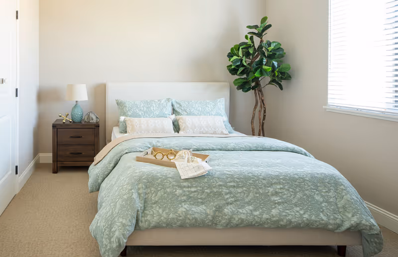 A neatly made bedroom with a bed, nightstand, lamp, and potted plant.