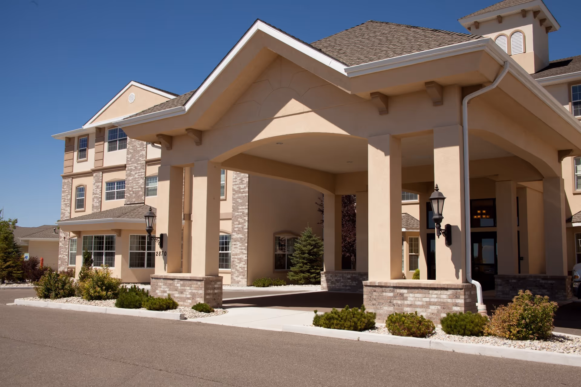 Exterior view of a senior living facility named Highland Village of Elko, showing the main entrance with a covered driveway supported by large columns, beige walls with brick accents, and landscaped bushes around the building under a clear blue sky.