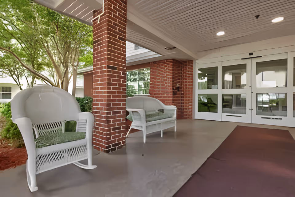 Covered outdoor seating area with white wicker chairs and a bench, brick pillars, and glass doors leading inside. Trees and greenery are visible in the background.