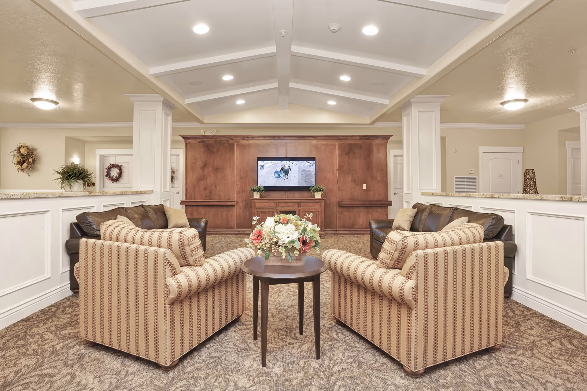 A cozy living room area in a senior living facility featuring two patterned armchairs and two dark leather sofas arranged around a small round wooden table with a floral centerpiece. A flat-screen TV is mounted on a wooden panel wall in the background. The room has beige walls, carpeted flooring, and recessed ceiling lights.
