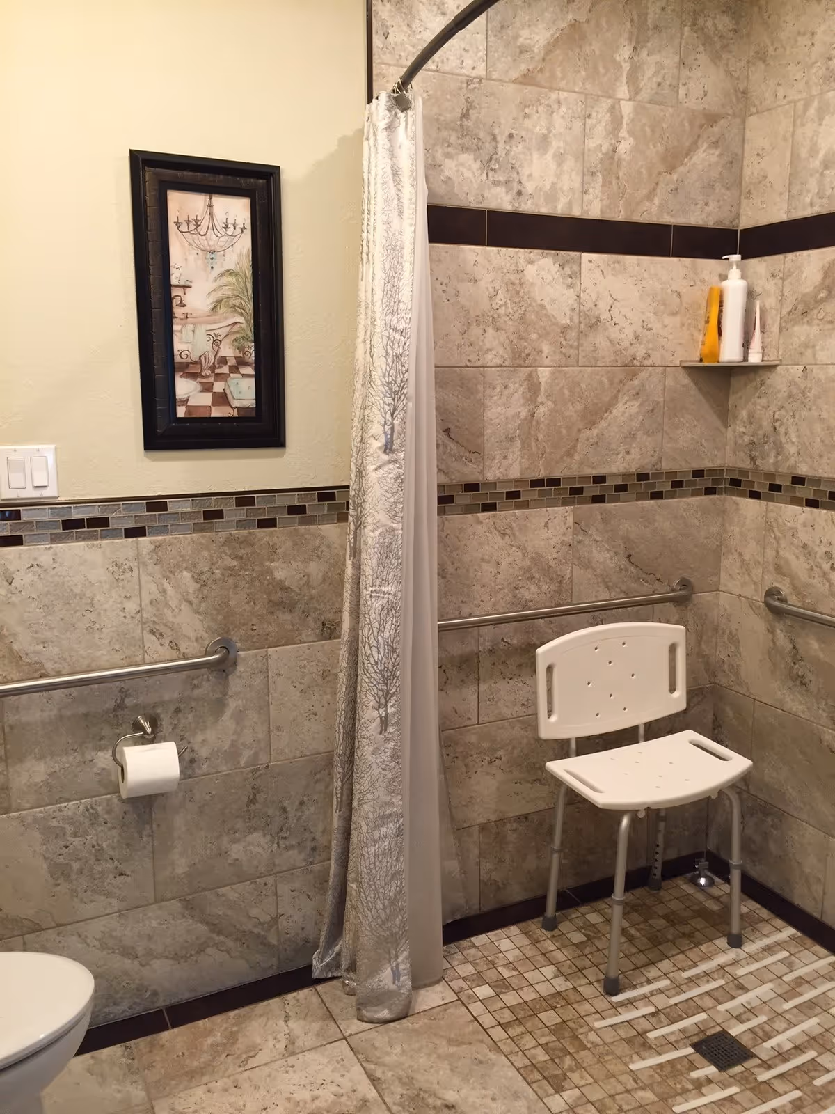 Bathroom with beige and brown tiled walls and floor. There is a white shower chair inside a walk-in shower area with a curtain partially drawn. The shower has grab bars on the walls and a small shelf holding bottles. A toilet paper holder with a roll is mounted on the wall outside the shower. A framed picture hangs on the wall above the toilet paper holder.