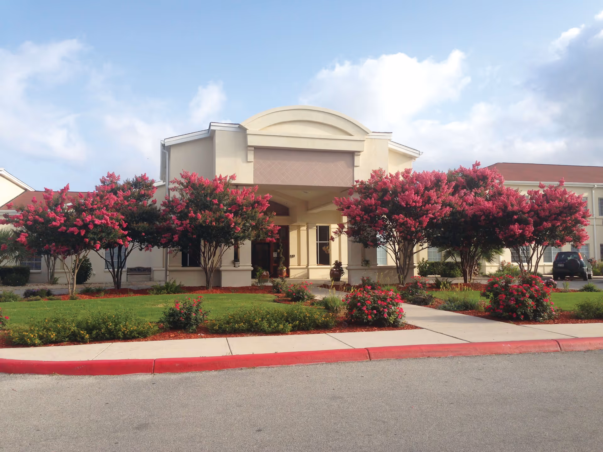 Front exterior view of a senior living facility named Esplanade Gardens with a covered entrance, surrounded by green lawns and blooming pink flowering trees under a partly cloudy sky.