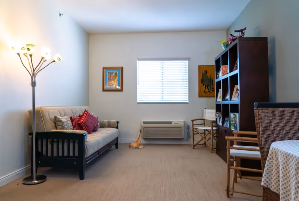A cozy living room with a beige futon sofa adorned with red and beige pillows on the left, a tall floor lamp with multiple lights beside it, a window with blinds in the center, and a dark wooden bookshelf filled with framed photos and decorative items on the right. There are two wooden chairs with white cushions and a table with a patterned tablecloth partially visible on the right side.