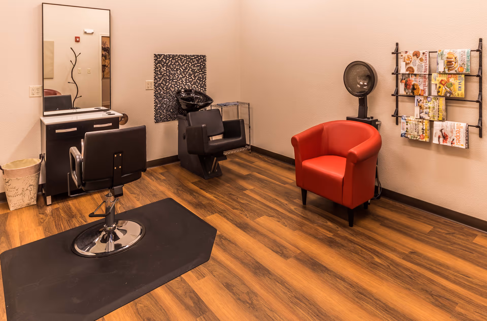 Interior of a salon room with a black styling chair in front of a mirror and counter, a black hair washing station with a chair, a red armchair, a hair dryer, and a wall-mounted magazine rack filled with magazines. The floor is wood and the walls are light-colored.