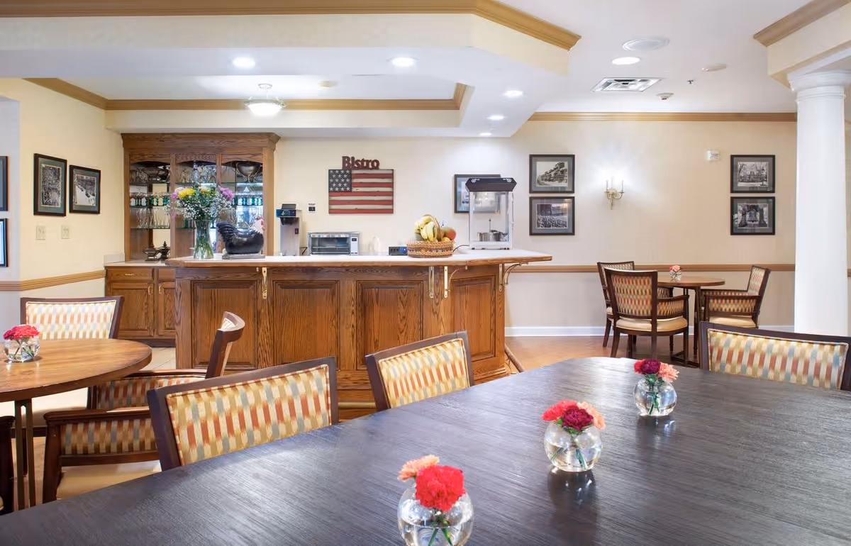 A cozy dining area in a senior living facility featuring wooden tables and chairs with patterned upholstery. The tables have small glass vases with red and pink flowers. In the background, there is a wooden counter with a basket of fruit, a popcorn machine, a toaster oven, and a coffee machine. The walls are decorated with framed black and white photos and an American flag with the word 'Bistro' above it. The room has warm lighting and a welcoming atmosphere.