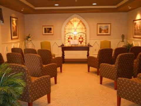 A small chapel or meditation room with rows of upholstered chairs facing a wooden table with a decorative stained glass window behind it. The room has soft lighting, framed artwork on the walls, and plants in the corners.
