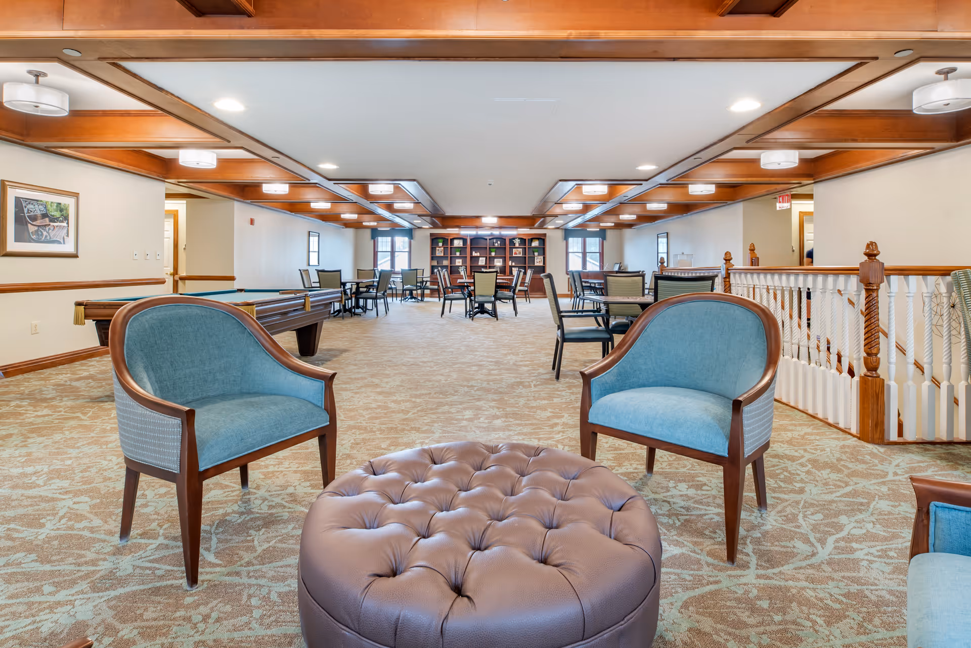 A spacious common area in a senior living facility featuring two blue upholstered chairs and a large round tufted ottoman in the foreground. The room has a patterned carpet, wooden ceiling beams with recessed lighting, several tables and chairs arranged throughout, a pool table on the left side, and a bookshelf with decorative items at the far end. The walls are light-colored with wooden trim, and there is a staircase with white spindles and wooden handrails on the right.