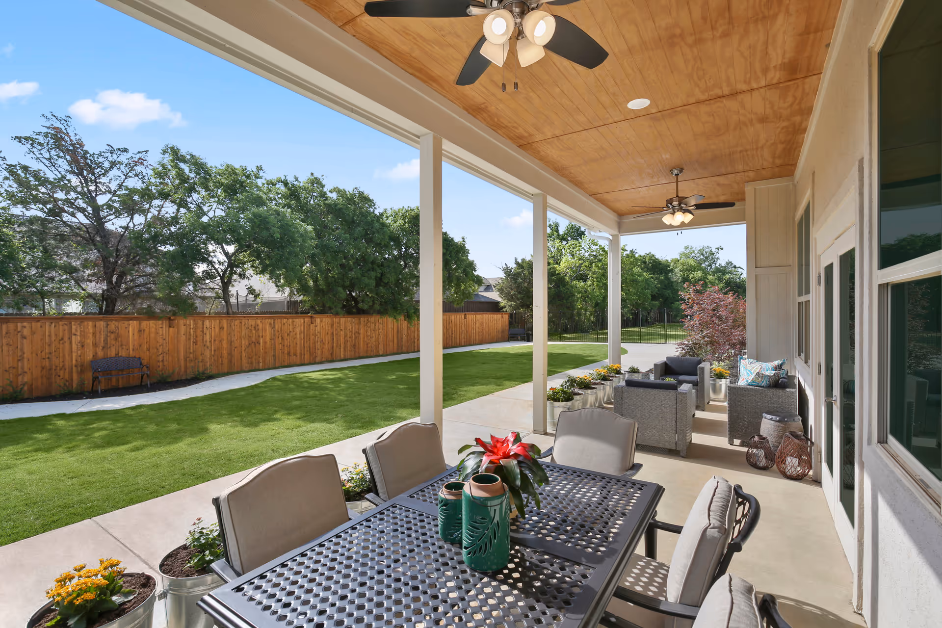 Covered outdoor patio area with a metal dining table and cushioned chairs, decorated with potted plants and a red flower centerpiece. The patio has a wooden ceiling with ceiling fans and overlooks a green lawn bordered by a wooden fence and trees. There is additional seating with cushioned armchairs and small tables along the patio.