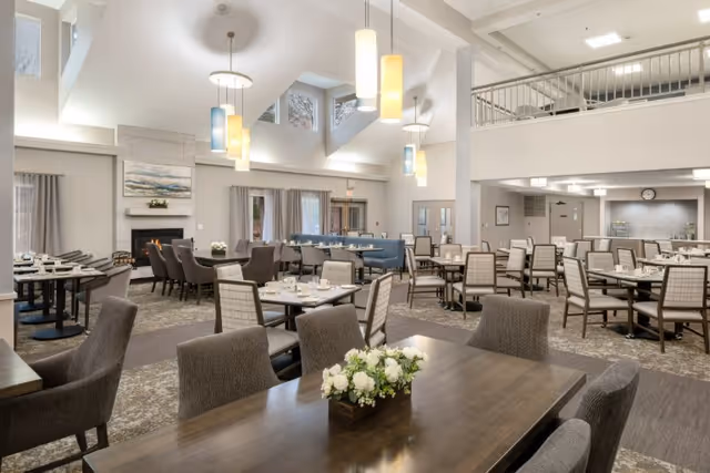 Spacious dining room in Bethel Park facility with multiple tables and chairs arranged neatly. The room features high vaulted ceilings with modern hanging pendant lights, large windows near the ceiling allowing natural light, a fireplace on one side, and a mix of cushioned and wooden chairs. The decor is neutral and elegant with floral centerpieces on the tables.