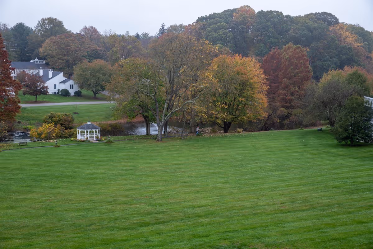 Wide view of a green lawn with a white gazebo, pond, and trees showing autumn foliage near a white building.