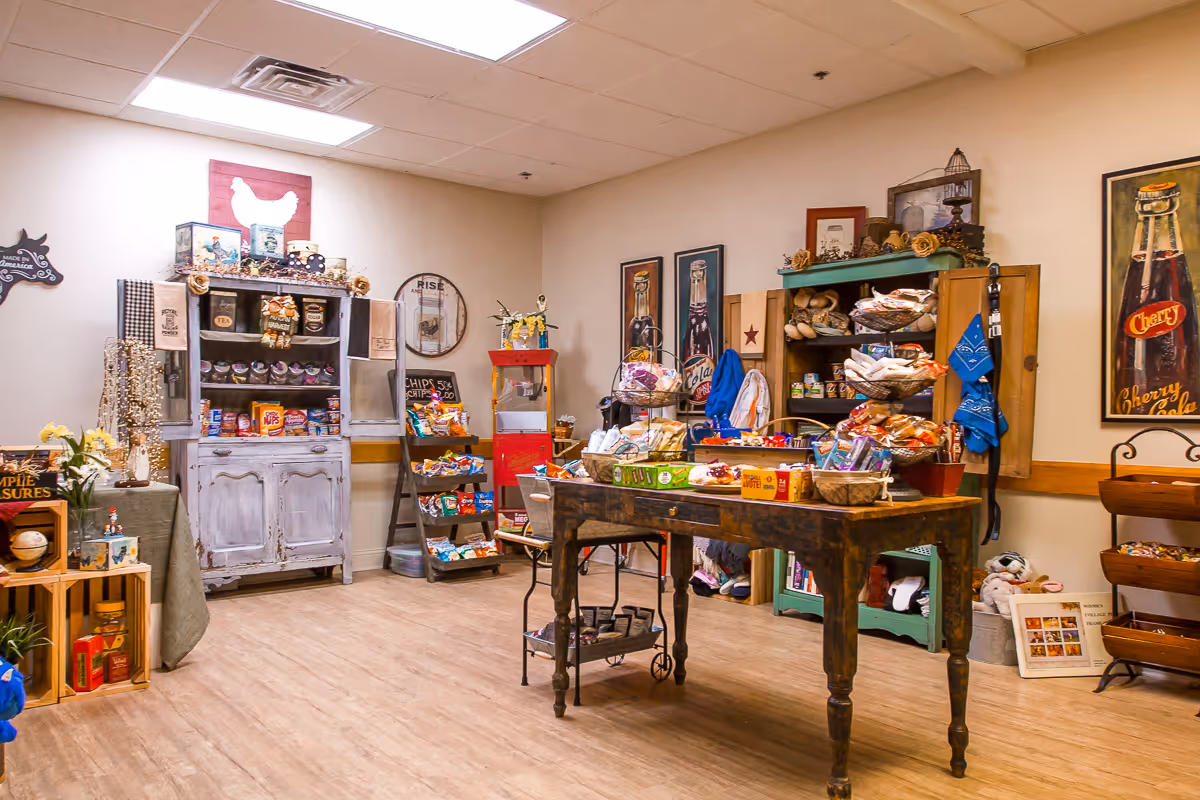 A cozy room with wooden flooring and warm lighting, featuring a rustic wooden table in the center filled with various packaged snacks and treats. Surrounding the table are vintage-style cabinets and shelves stocked with more snacks, decorative items, and framed artwork on the walls. The room has a welcoming, homey atmosphere.