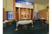 Interior view of a chapel or prayer room with an altar in the center, flanked by two candle holders. The room features stained glass windows on the brick walls and a green carpeted floor. There are benches and religious items arranged around the altar.