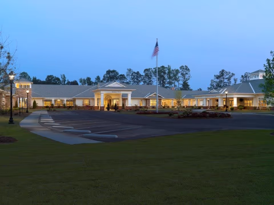 Exterior view of Wesley Pines senior living facility at dusk with a large parking lot, an American flag on a flagpole in front, and the building warmly lit from inside. Trees are visible in the background under a clear blue sky.